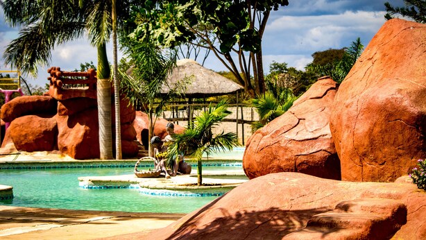 A pool area with large rock formations, a thatched-roof structure, and palm trees. - Dream Valley Park & Lodge (Lusaka)