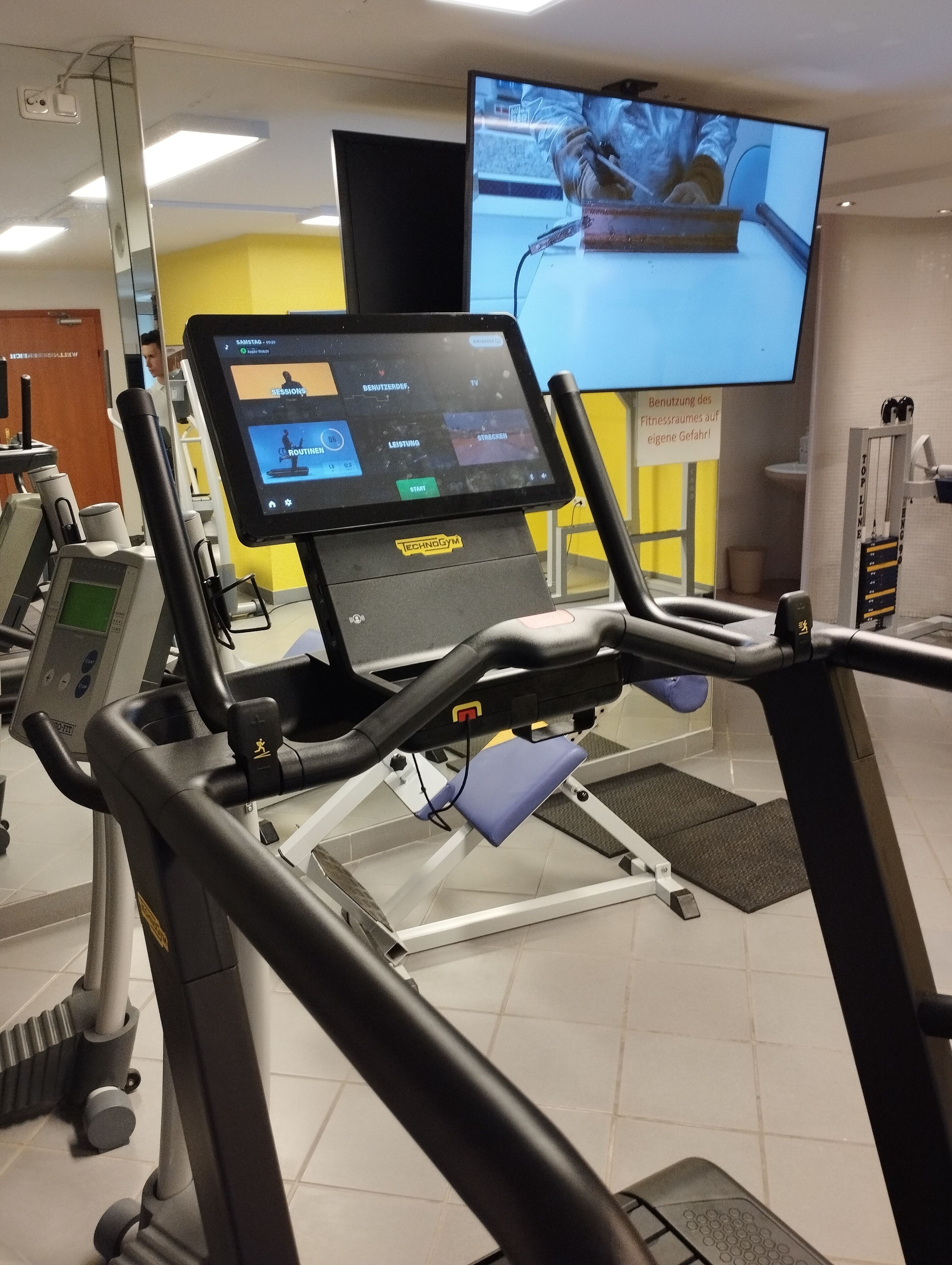 A gym with a treadmill, a digital screen, and a television displaying a workout.