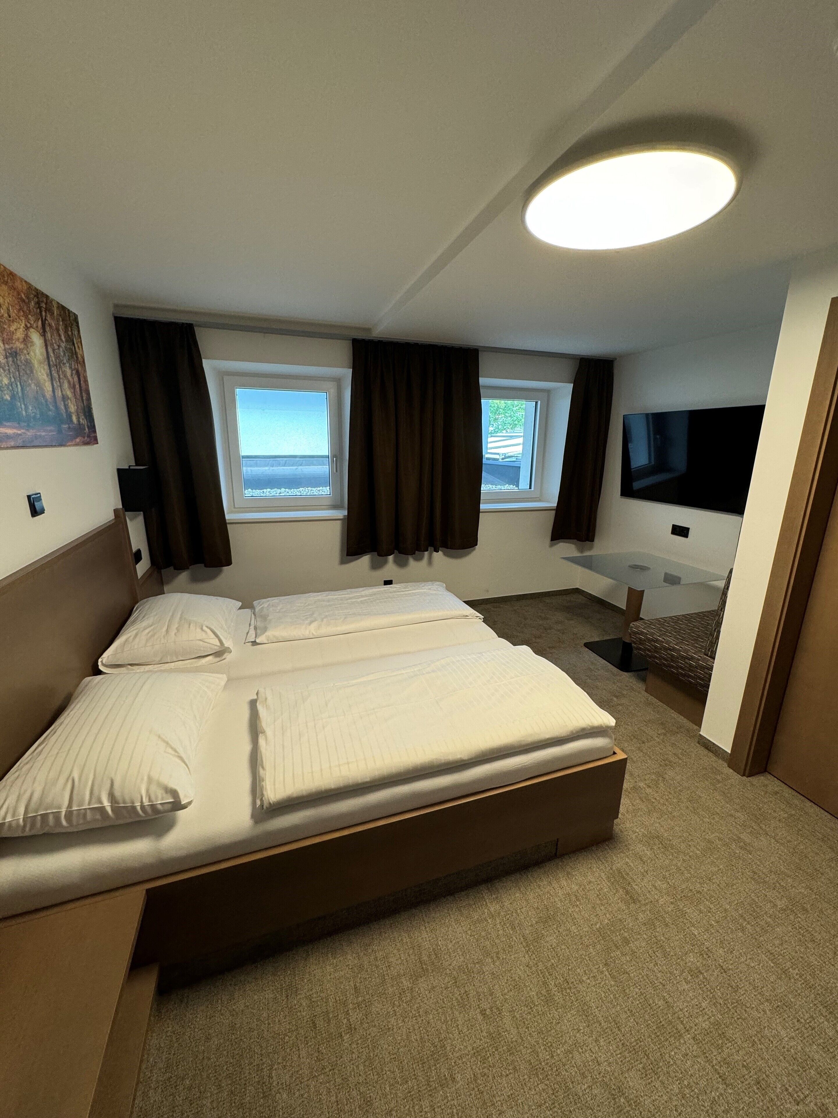 A hotel room with a large bed, two windows, a flat-screen TV, and a small desk area.
