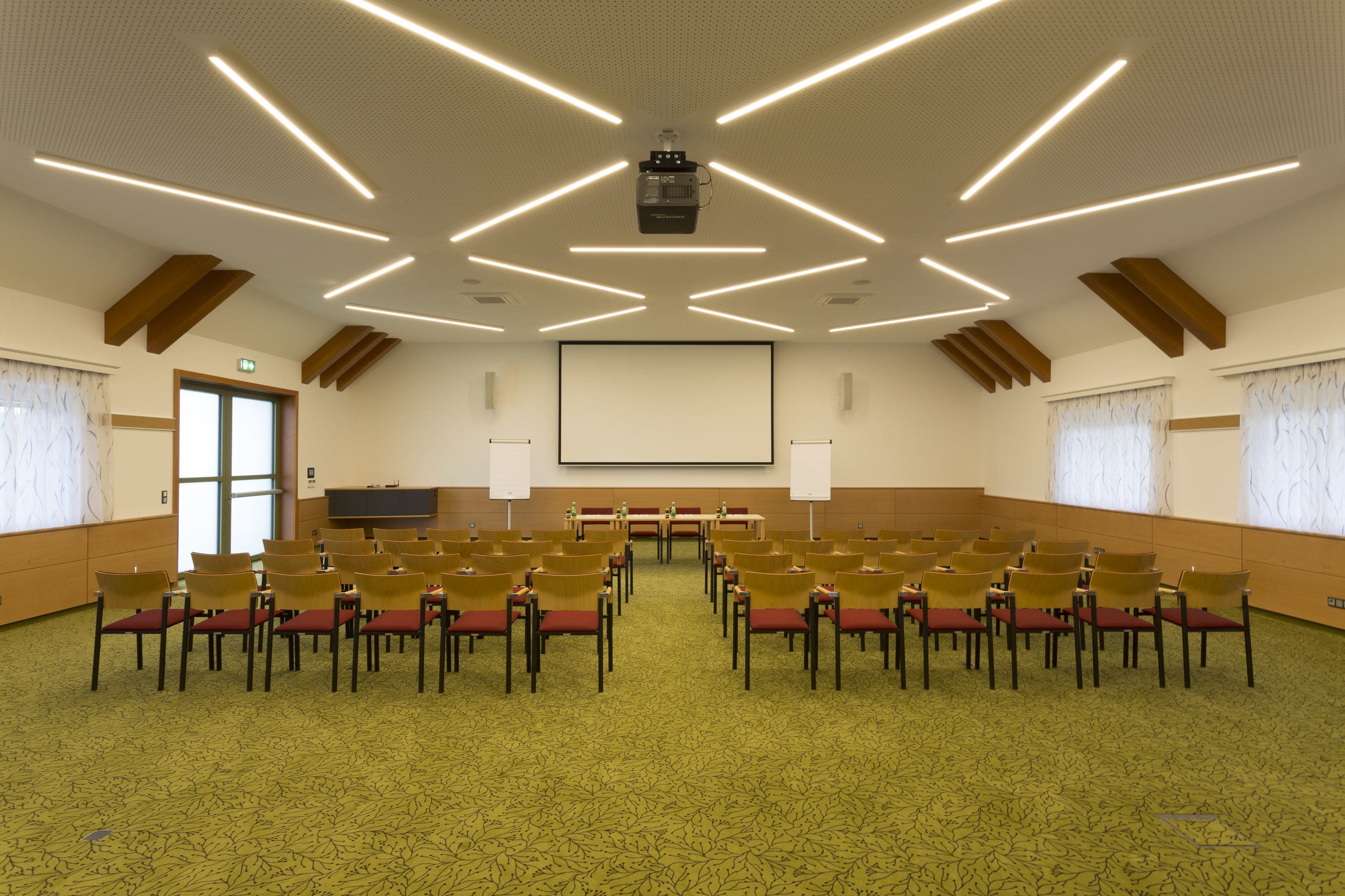 A conference room with a projector, a large screen, rows of chairs, and a carpeted floor.