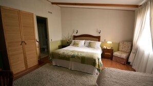 Quarto com cama, mesa de cabeceira, cadeira, guarda-roupa de madeira e porta para outro espaço.