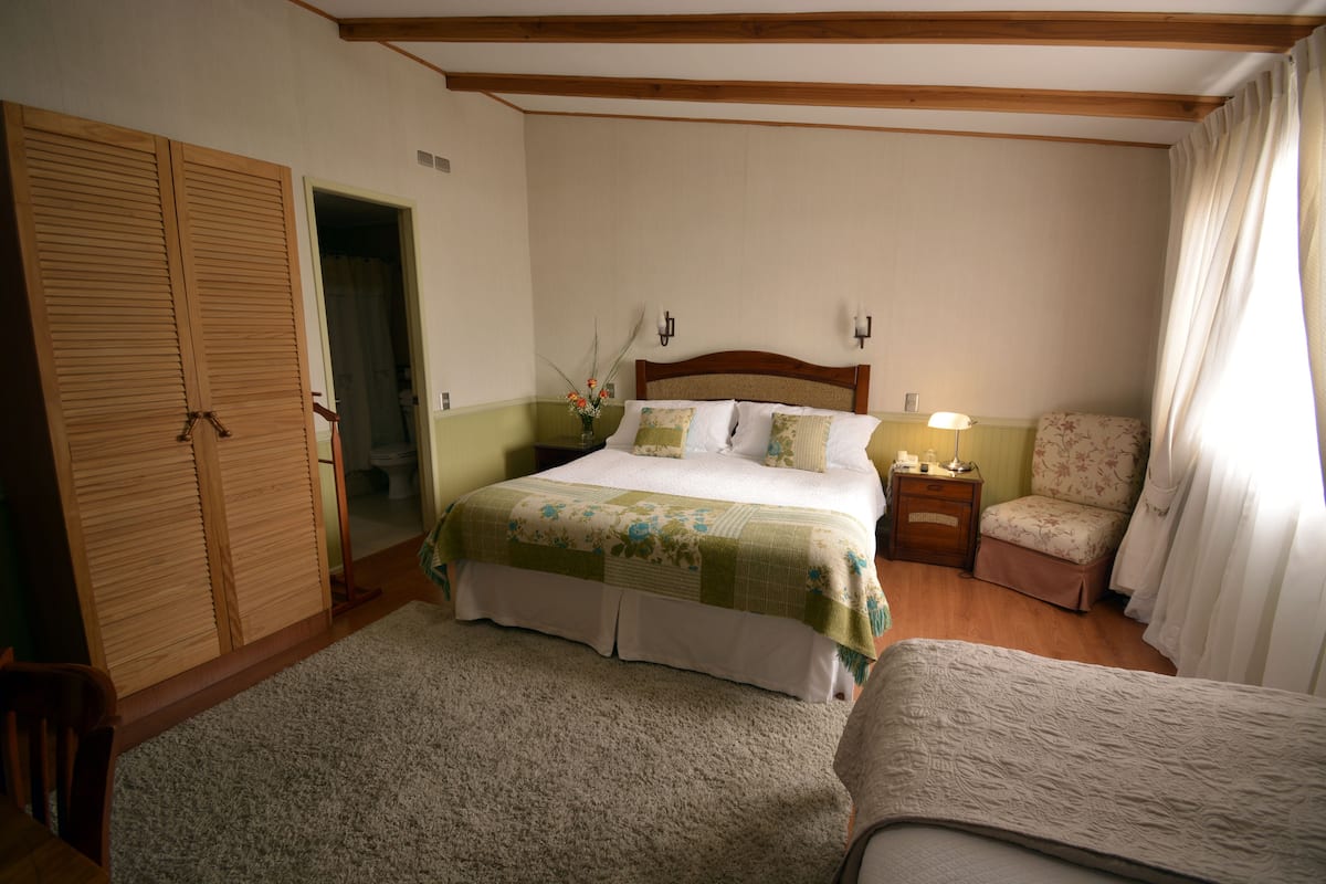 A bedroom with a bed, a nightstand, a chair, a wooden wardrobe, and a door leading to another room.