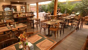 Restaurante com várias mesas e cadeiras de madeira, um balcão com vitrine e vista para o exterior arborizado.