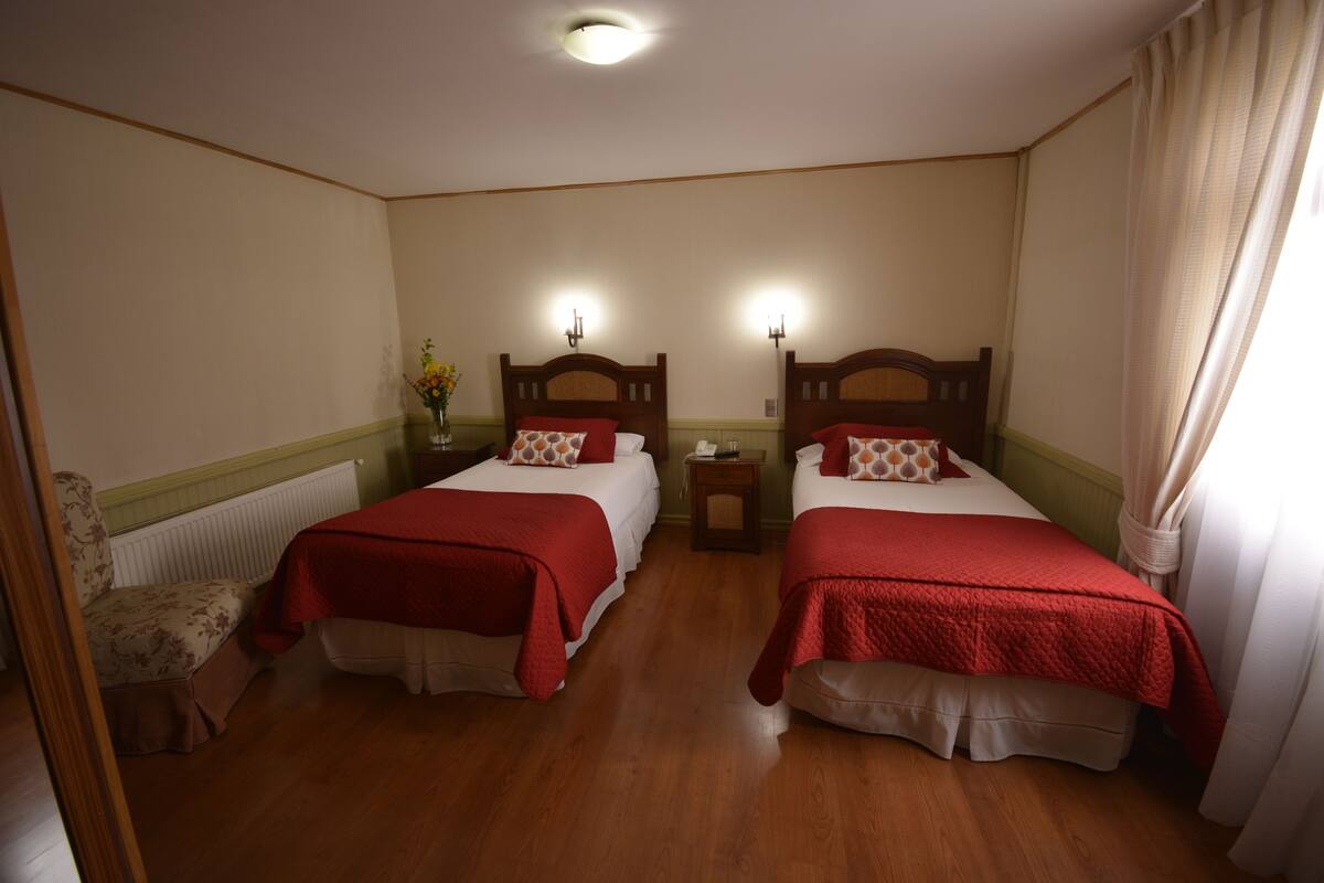 A hotel room with two beds, wooden headboards, a nightstand, a chair, and a radiator.