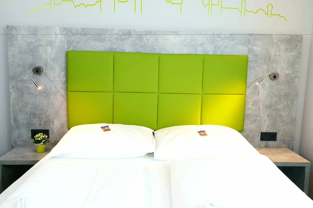 Un dormitorio con cabecera acolchada verde, ropa de cama blanca y una lámpara fijada a la pared.
