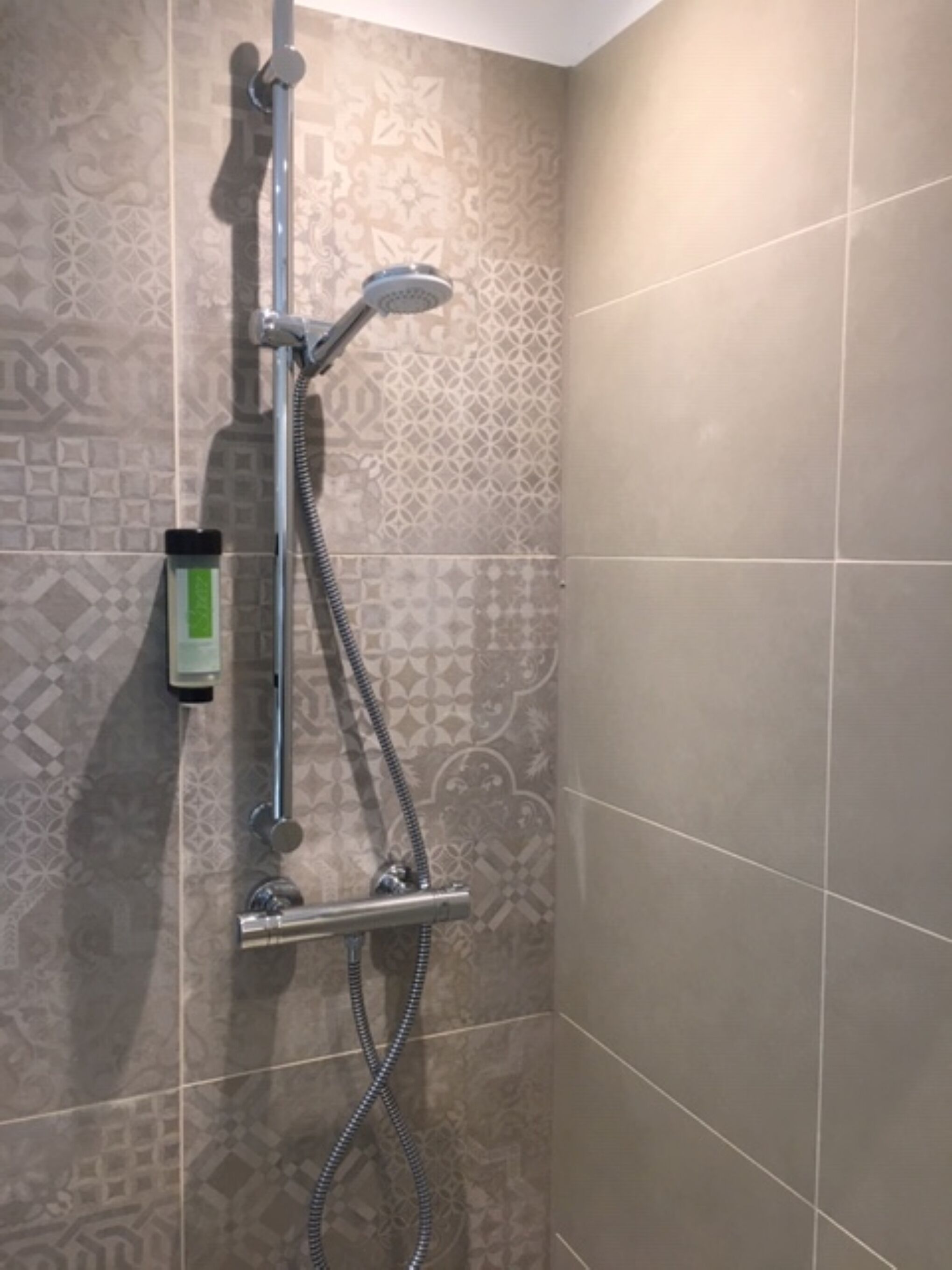 A shower with a handheld showerhead and a handheld hose, mounted on a wall with patterned tiles.