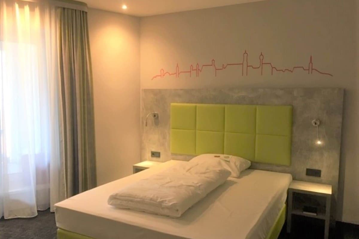 Une chambre d’hôtel comprenant un lit, une tête de lit, une table de chevet, une commode et une fenêtre avec des rideaux.