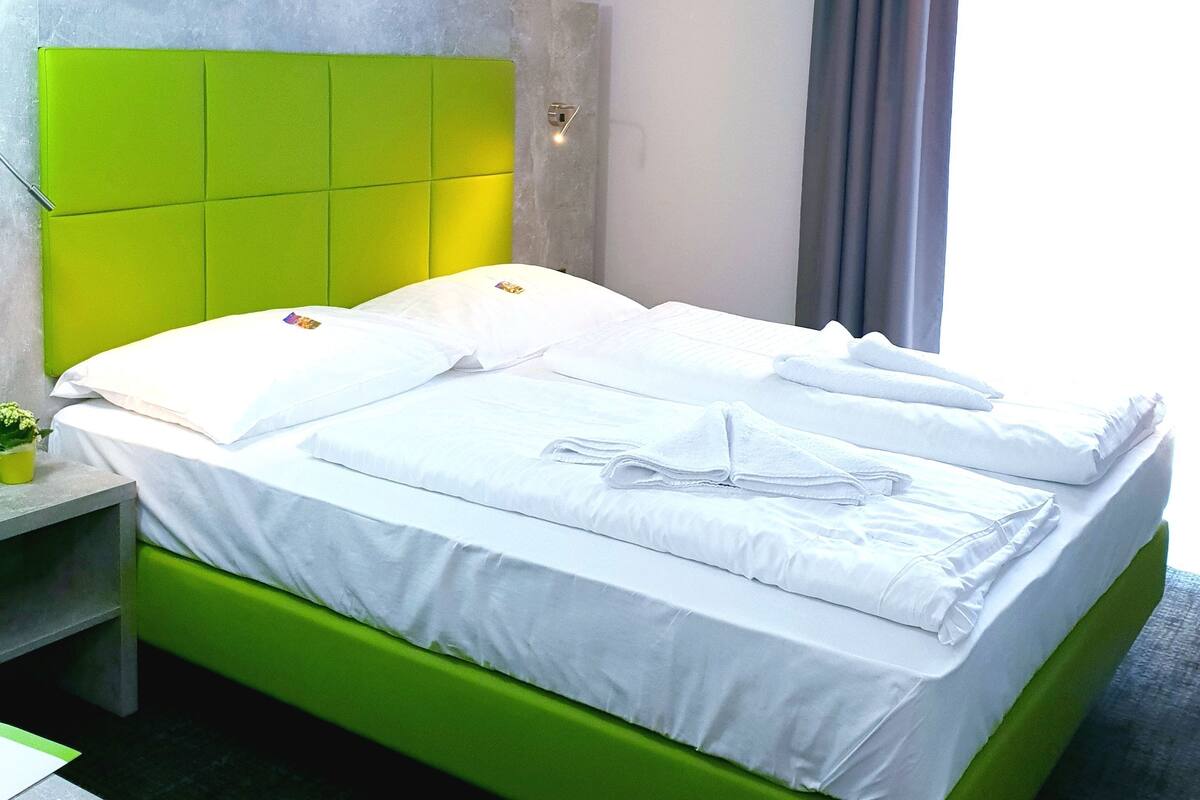 Habitación de hotel con cabecera verde, cama con ropa de cama blanca, mesita de noche con una planta y una pared con diseño de línea clara.