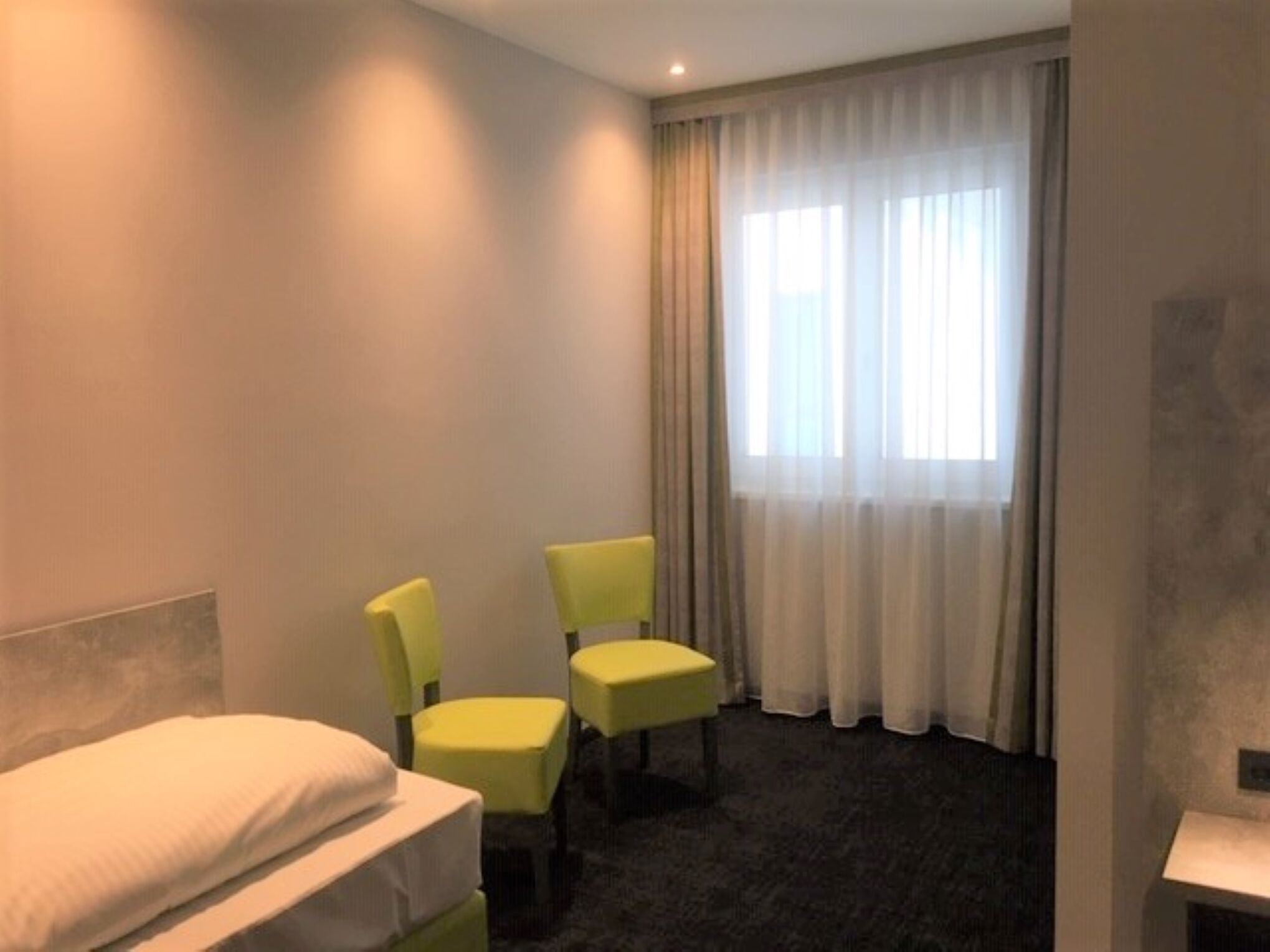 Ein Hotelzimmer mit einem Bett, zwei gelben Stühlen, einem Fenster mit durchsichtigen Vorhängen und einem Teppichboden.