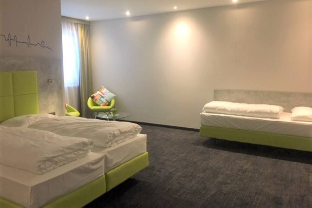 Una habitación de hotel con dos camas, un cabecero verde, una mesita redonda pequeña con una silla, y una pared con un dibujo lineal sencillo.