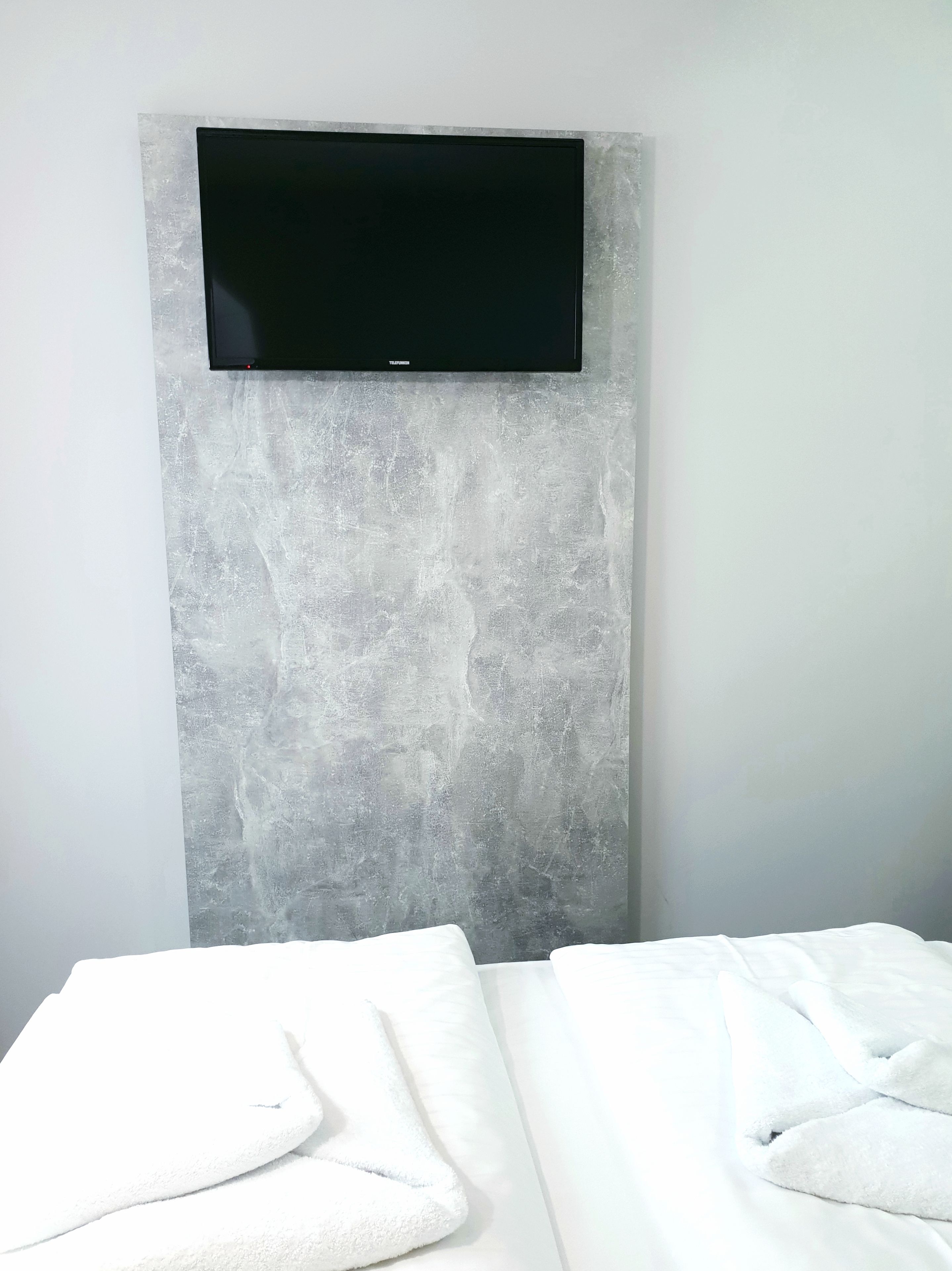 Une chambre à coucher moderne avec un téléviseur à écran plat fixé à un mur gris texturé, un lit soigneusement fait avec du linge de lit blanc, et un design épuré et minimaliste.