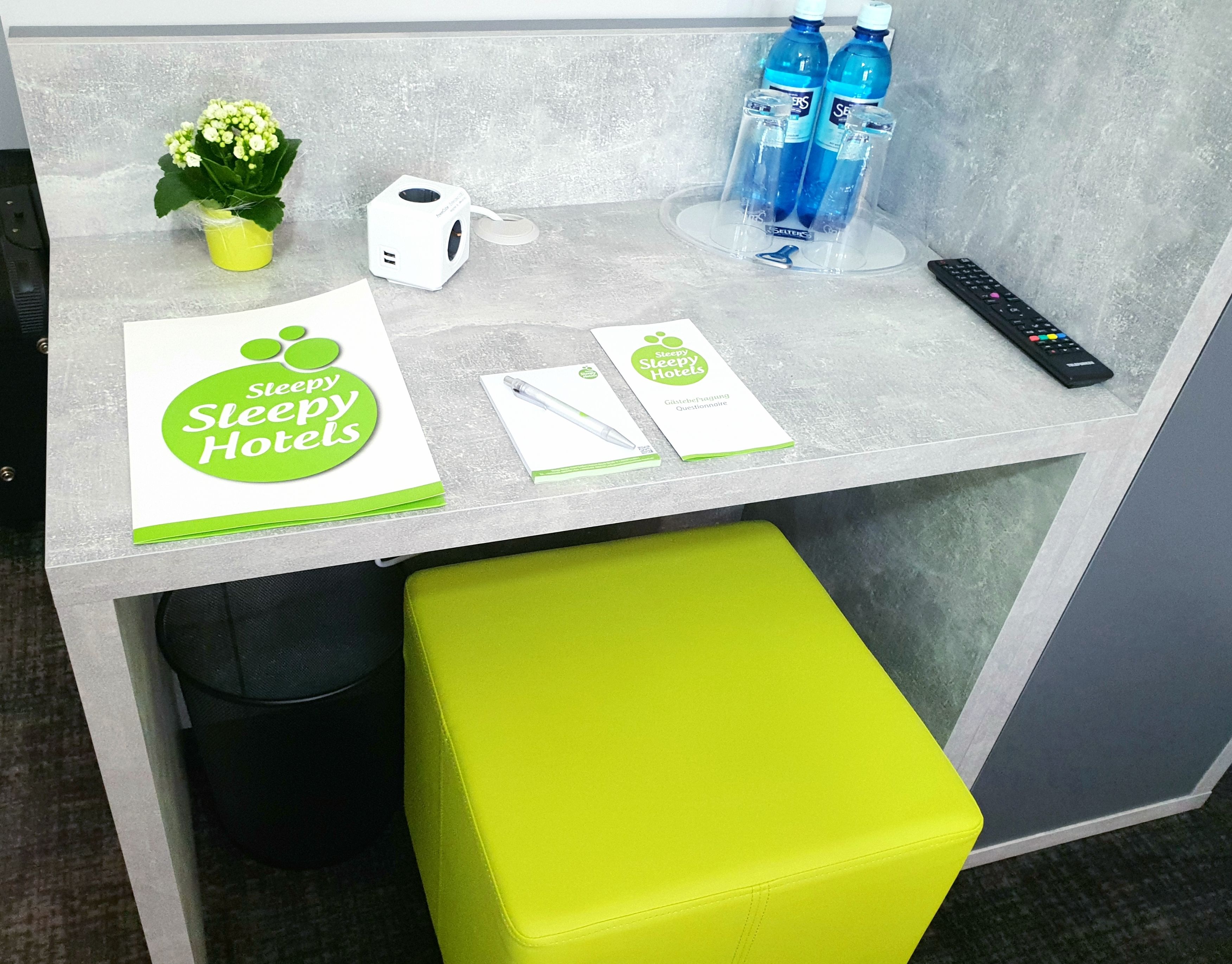 Un mostrador de recepción con un logotipo verde y blanco, una pequeña planta en maceta, un altavoz blanco, botellas de agua azules, un control remoto y un cuestionario.