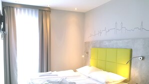 A hotel room with a bed, a headboard, a wall with a city skyline design, and a window with curtains. - SleepySleepy Hotel Dillingen (Dillingen an der Donau)