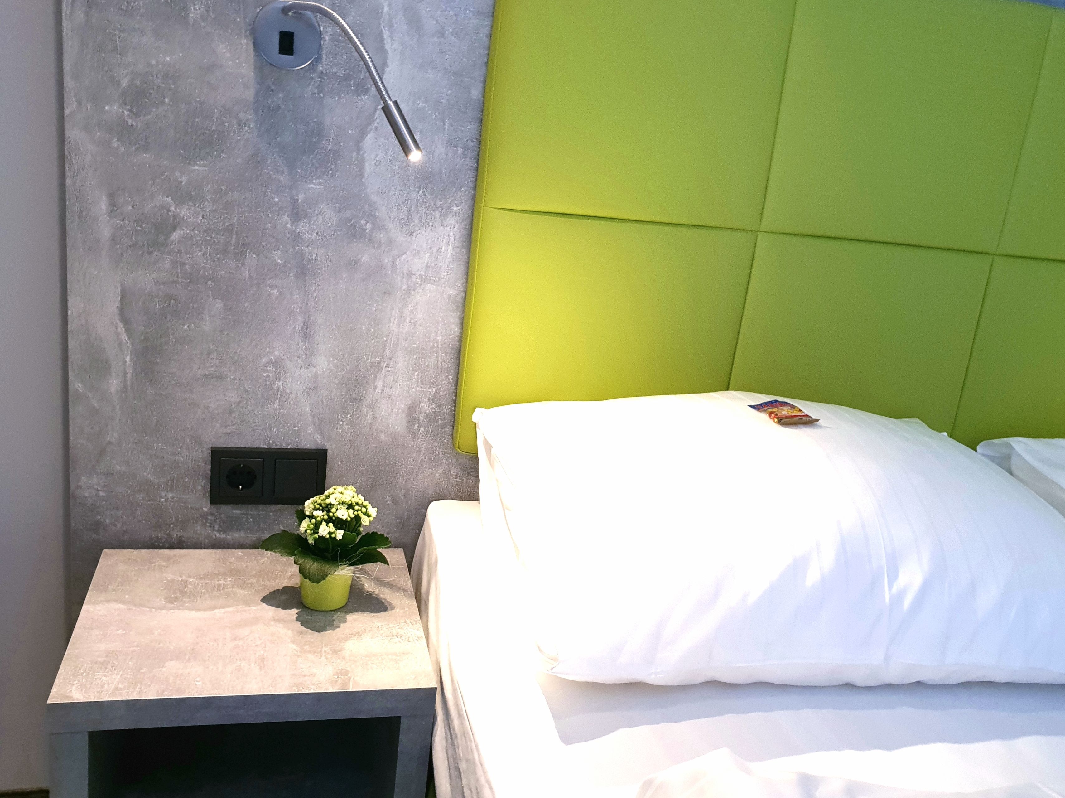 Una habitación de hotel moderna con una pared gris con textura, un cabecero verde, una mesita de noche con una planta en maceta y una cama bien hecha con ropa blanca.