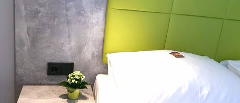 Una habitación de hotel moderna con una pared gris con textura, un cabecero verde, una mesita de noche con una planta en maceta y una cama bien hecha con ropa blanca.