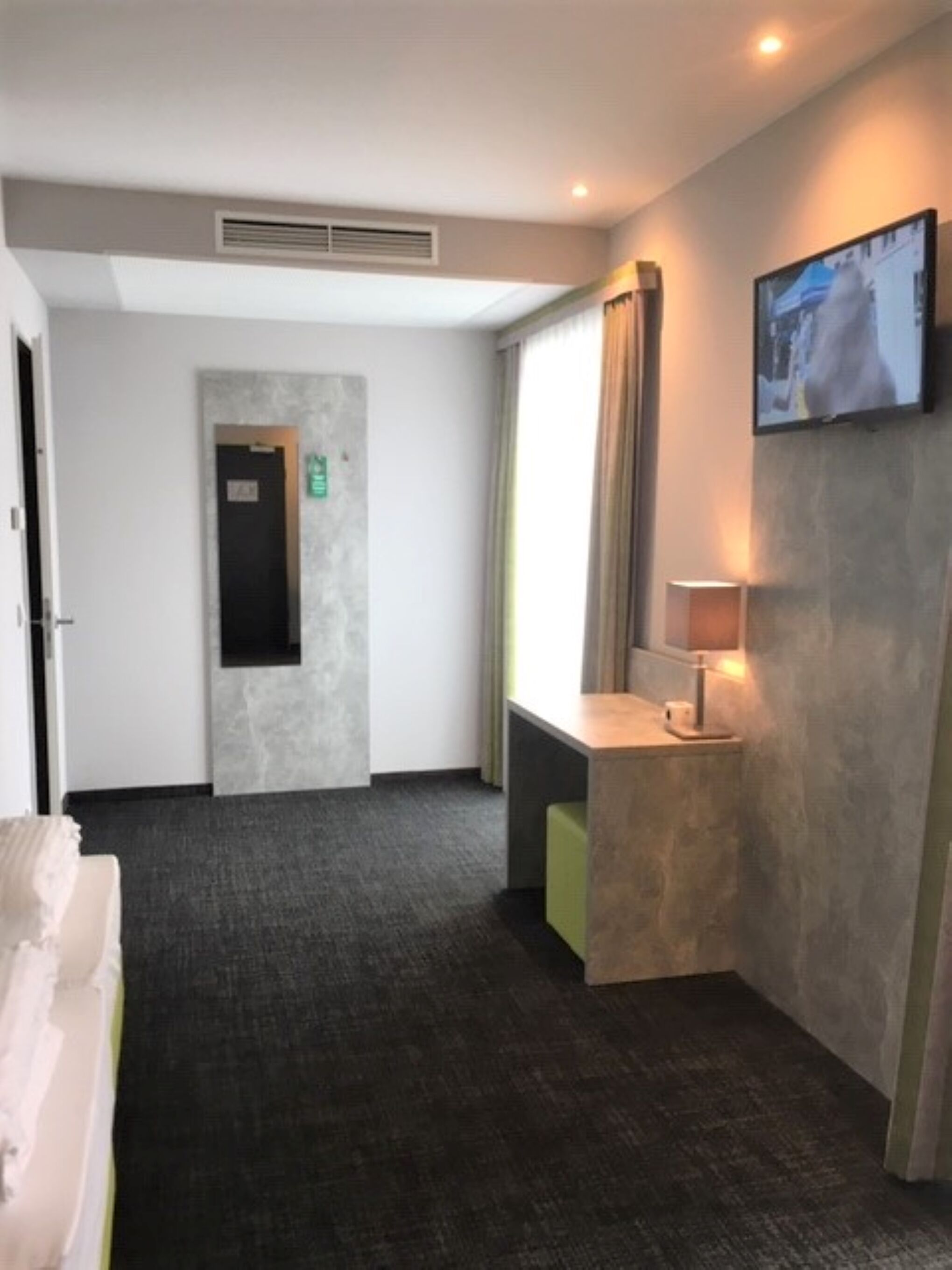 A hotel room with a dark carpet, a bed, a small desk with a lamp, a television mounted on the wall, and a door with an emergency exit sign.