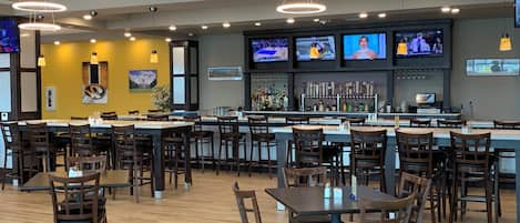 A restaurant with multiple tables and chairs, a bar area with a variety of bottles, and several televisions mounted on the wall.