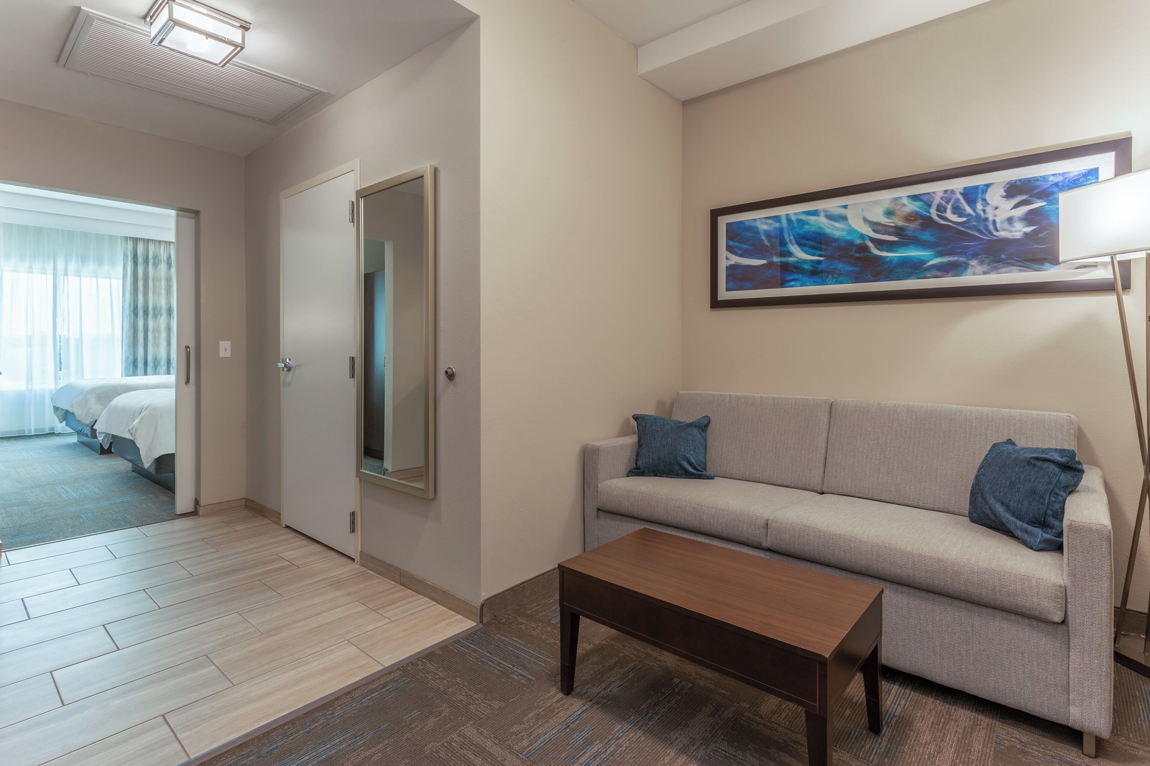 Ein Hotelzimmer mit einer Couch, einem Couchtisch, einem Bild an der Wand und Blick auf das angrenzende Zimmer.