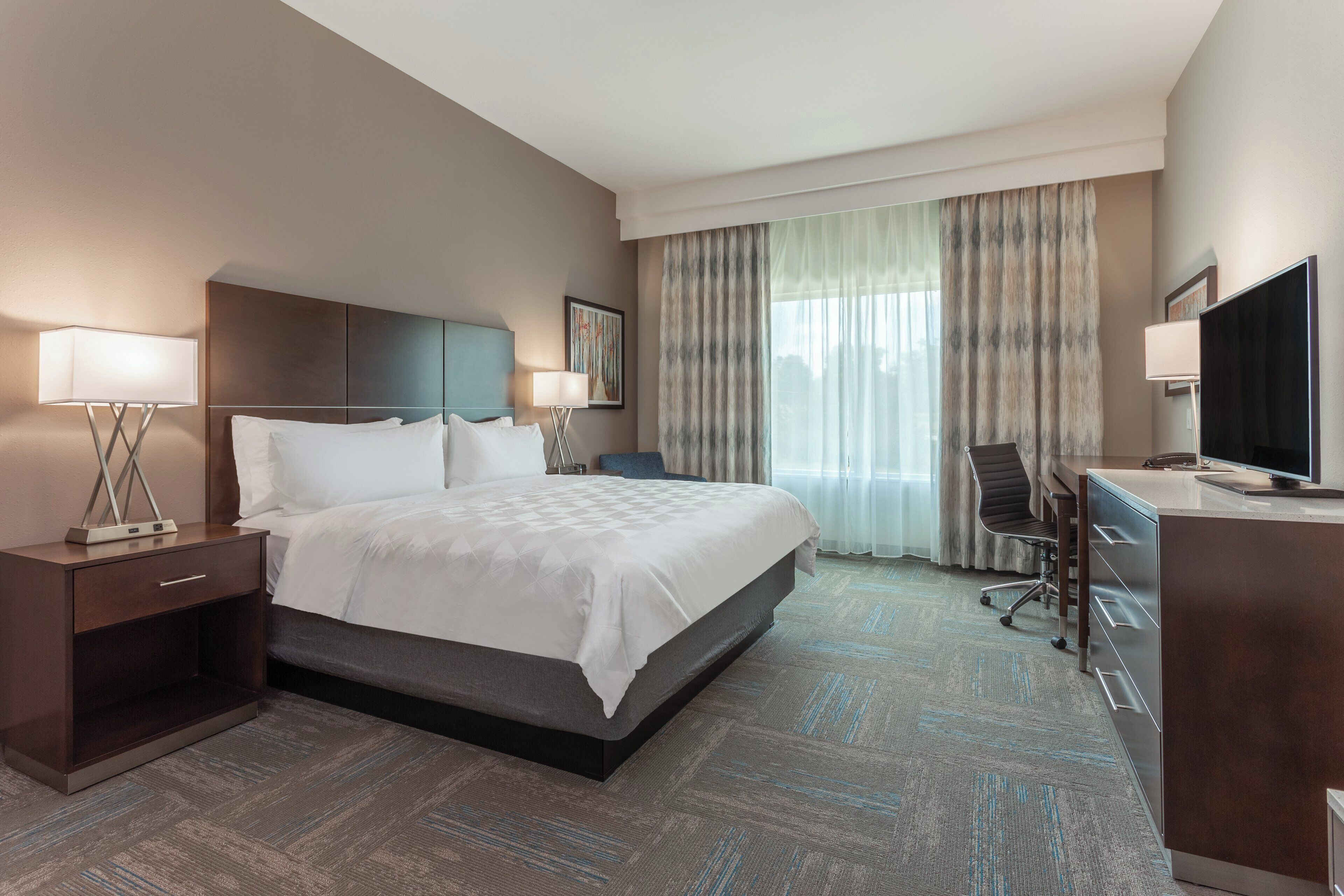 A hotel room with a bed, two bedside lamps, a desk with a chair, a television, and a large window with curtains.