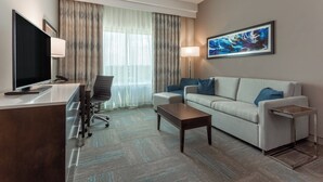 Een moderne hotelkamer met een flatscreen televisie, een bureau met lades, een bank, een salontafel en een groot raam met gordijnen.
