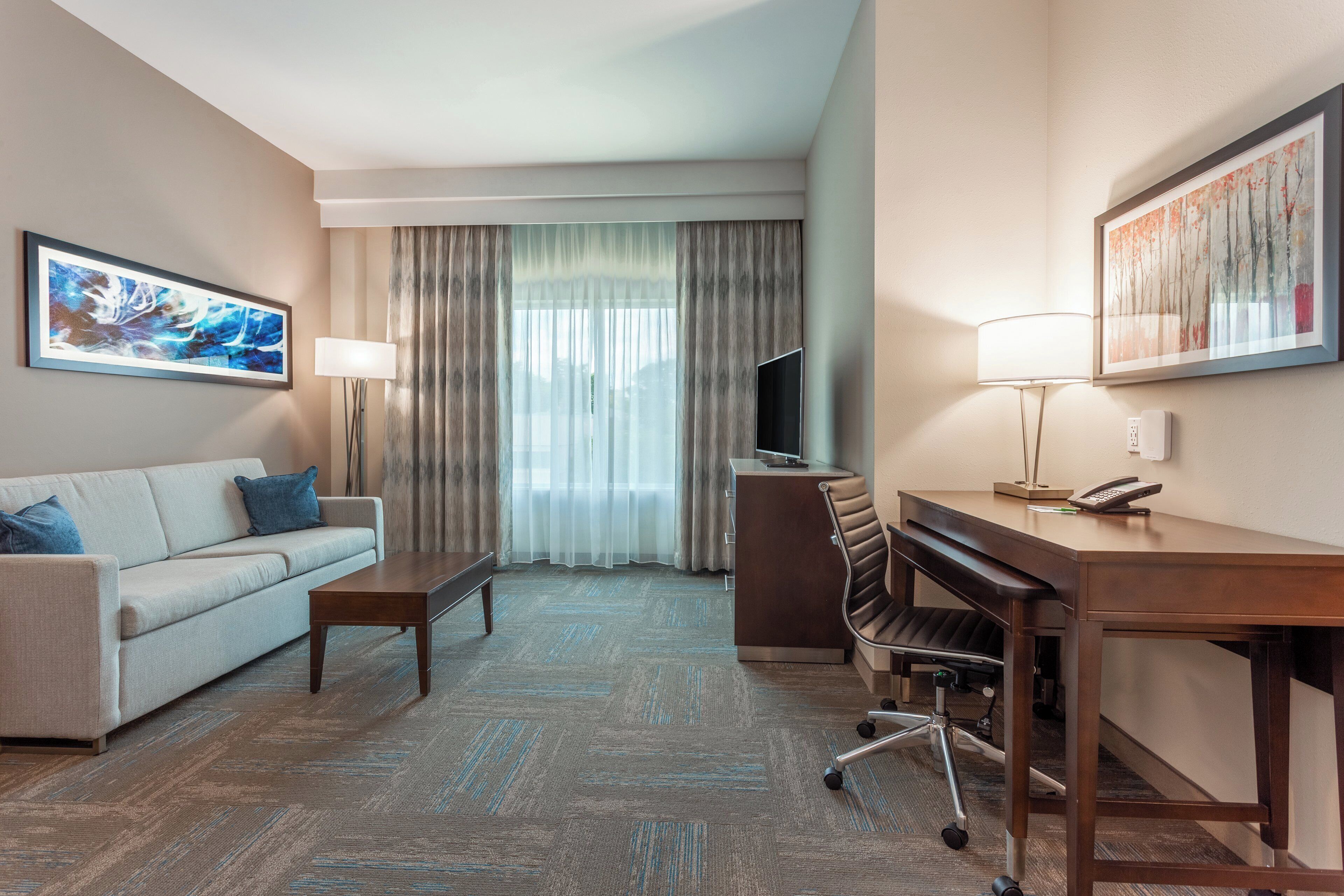 A hotel room with a sofa, a desk, a chair, a TV, and a painting on the wall.
