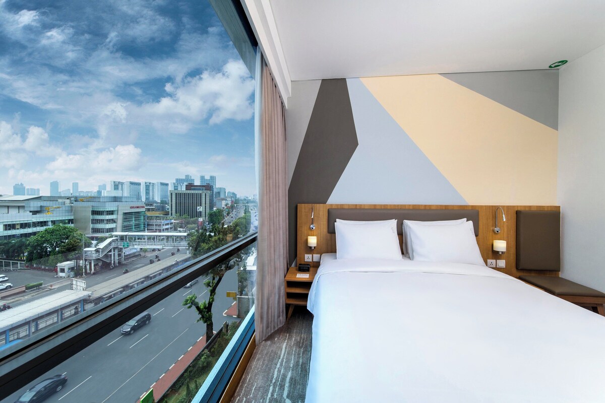 Une chambre d’hôtel avec un lit, des tables de chevet et une vue sur le paysage urbain, visible à travers une grande fenêtre.
