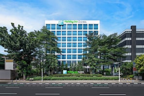 A Holiday Inn Express hotel with a modern design, featuring a prominent logo, large windows, and a landscaped front area with trees and a bench. - Holiday Inn Express Jakarta Menteng by IHG (Jakarta)