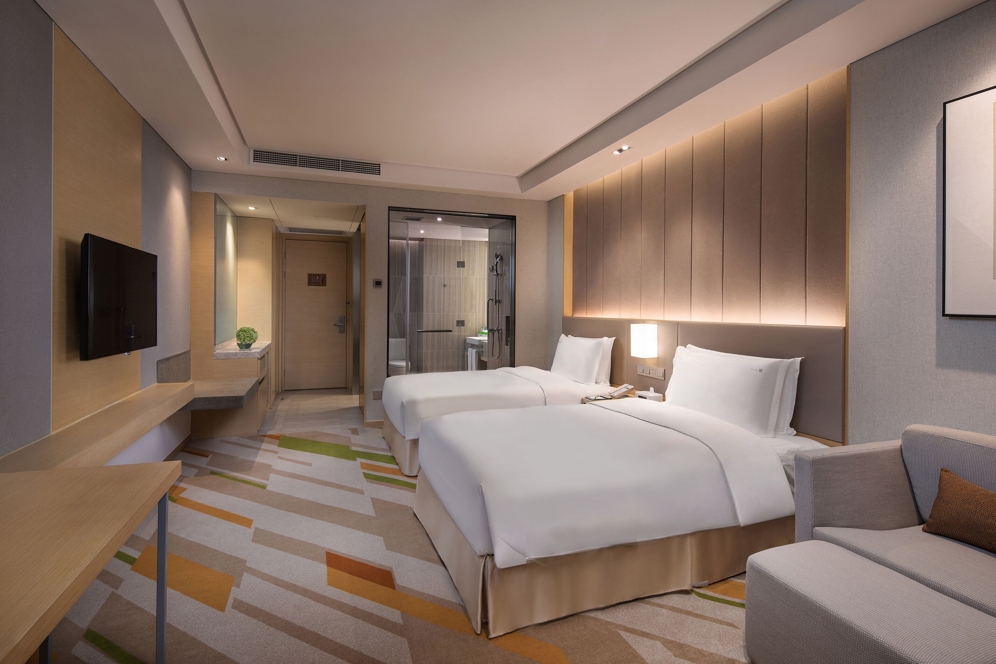 A hotel room with two beds, a sofa, a TV, and a bathroom visible through a glass door.