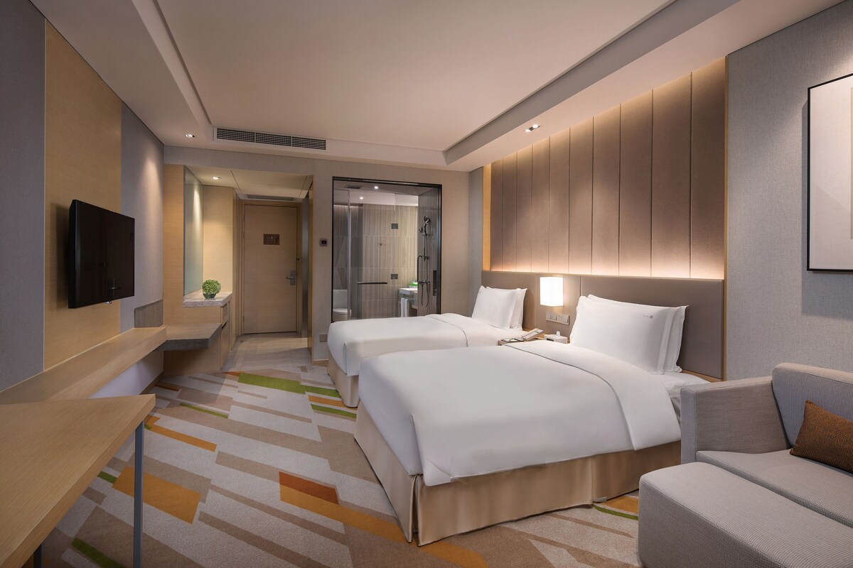 A hotel room with two beds, a sofa, a TV, and a bathroom visible through a glass door.