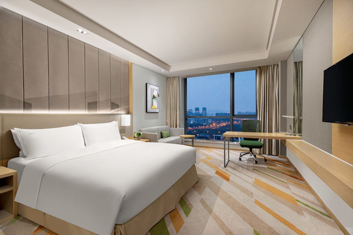 Ein Hotelzimmer mit einem großen Bett, einem Schreibtisch, einem Sessel, einem Fernseher und Blick auf die Stadt.