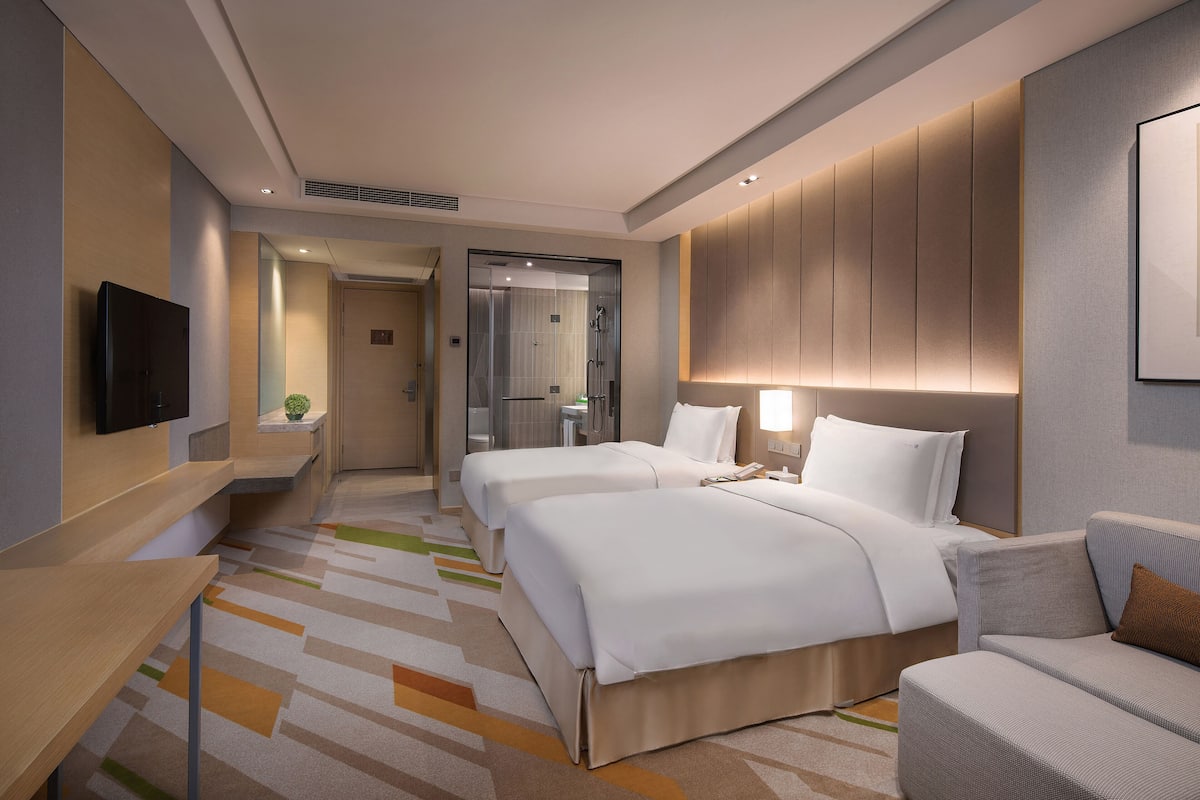 Ein Hotelzimmer mit zwei Betten, einem Sofa, einem Fernseher und einem durch eine Glastür einsehbaren Badezimmer.