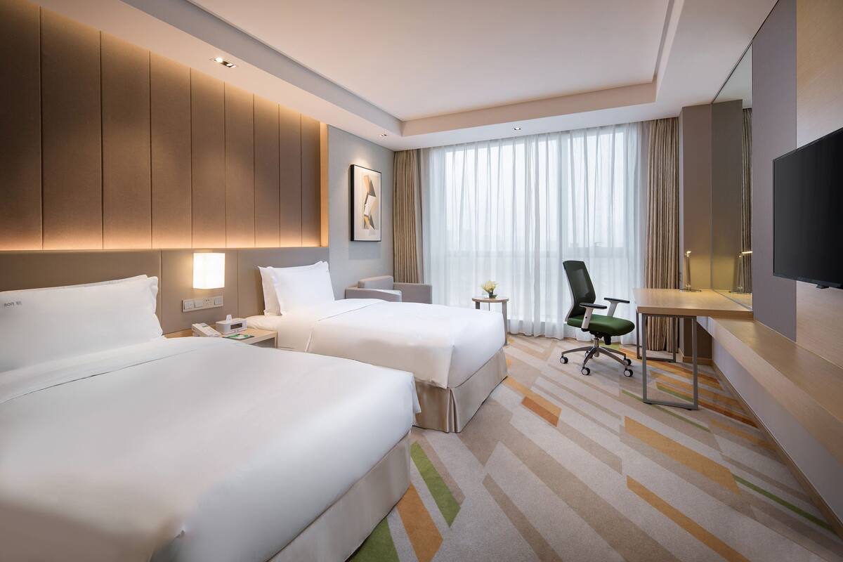 Ein Hotelzimmer mit zwei Betten, einem Schreibtisch, einem Sessel, einem Fernseher und einem großen Fenster mit Vorhängen.