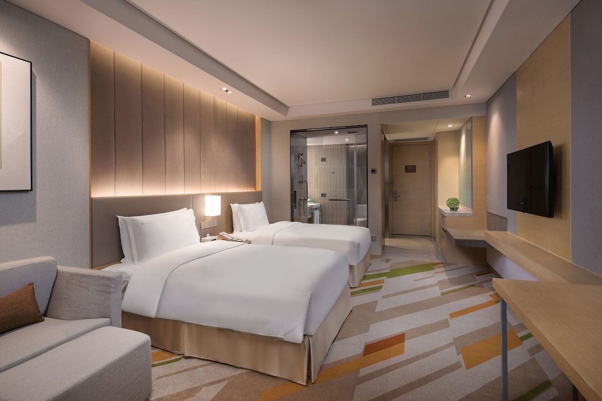 Ein modernes Hotelzimmer mit zwei Betten, einem Sofa, einem Fernseher und einem durch eine Glastür sichtbaren Badezimmer.