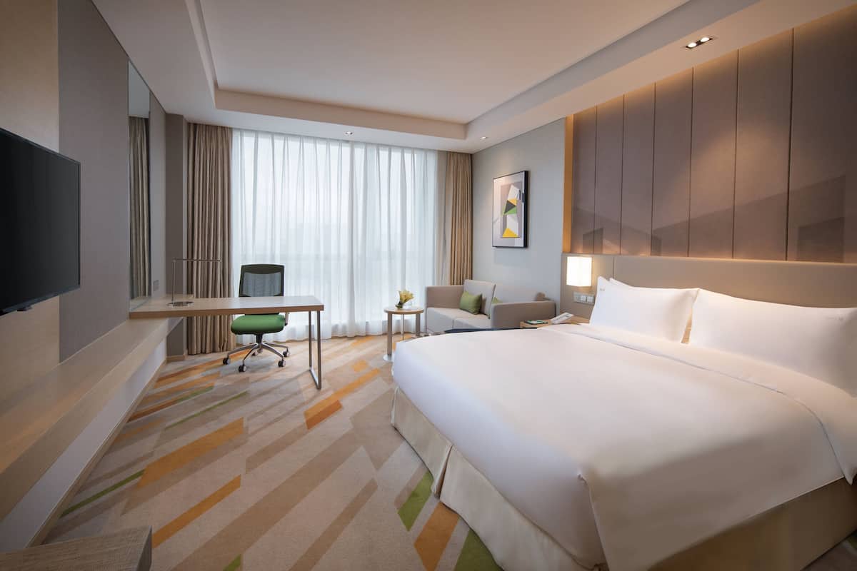 Ein Hotelzimmer mit einem großen Bett, einem Schreibtisch mit Stuhl, einem Fernseher und einem Fenster mit Vorhängen.