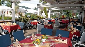 Een terrasrestaurant met tafels met rode tafelkleden, blauwe stoelen en bloemstukken.