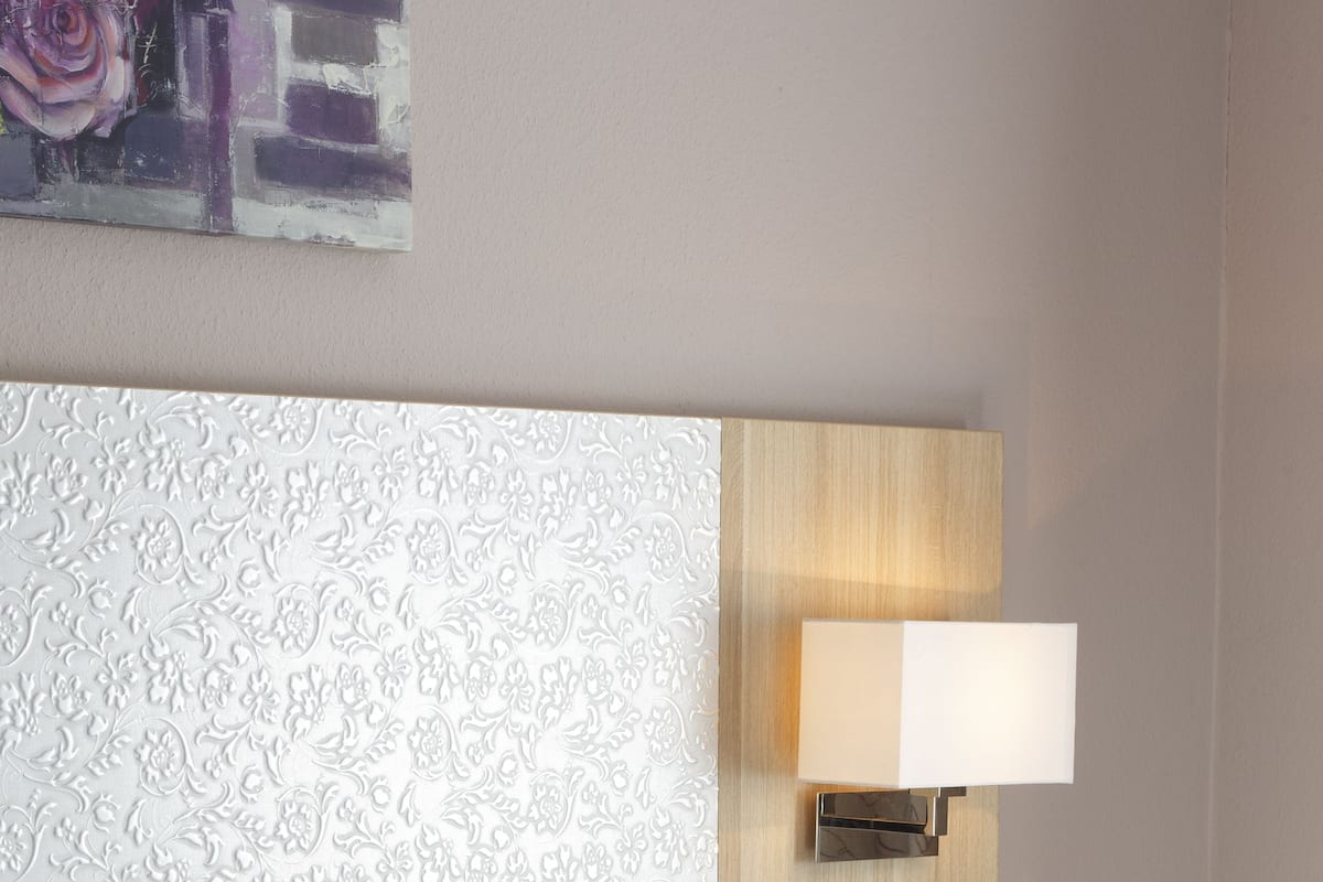 Een hotelkamer met een bed, witte kussens, een hoofdbord met een structuur en een wandlamp.