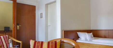 Una habitación de hotel con una cama y una silla de madera, una mesita con una botella y un vaso, y un televisor sobre un mueble.
