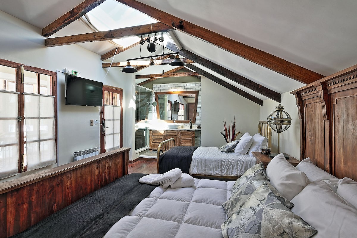 A bedroom with a sloped ceiling, wooden beams, a large bed with pillows, a bedside table, and a mirror.