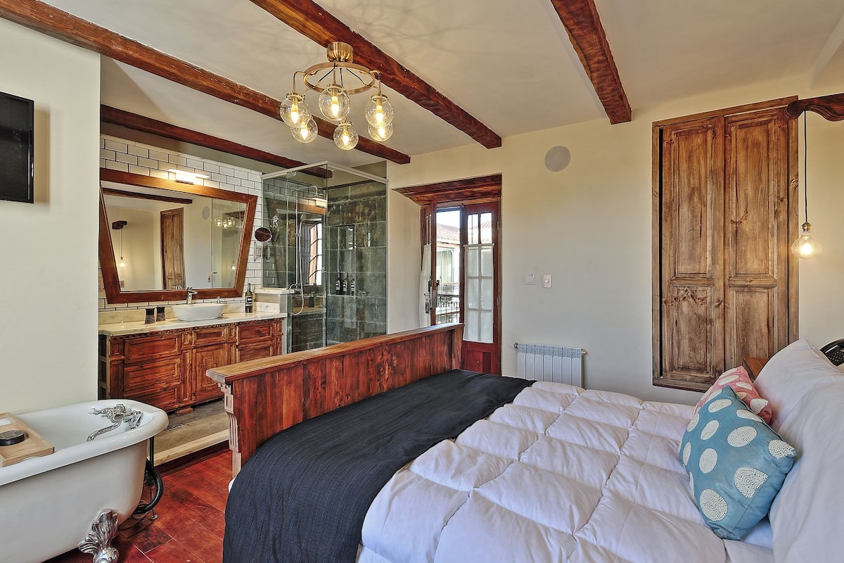 A bedroom with a large bed, wooden beams, a wooden wardrobe, a mirror, a bathroom with a bathtub, and a chandelier.