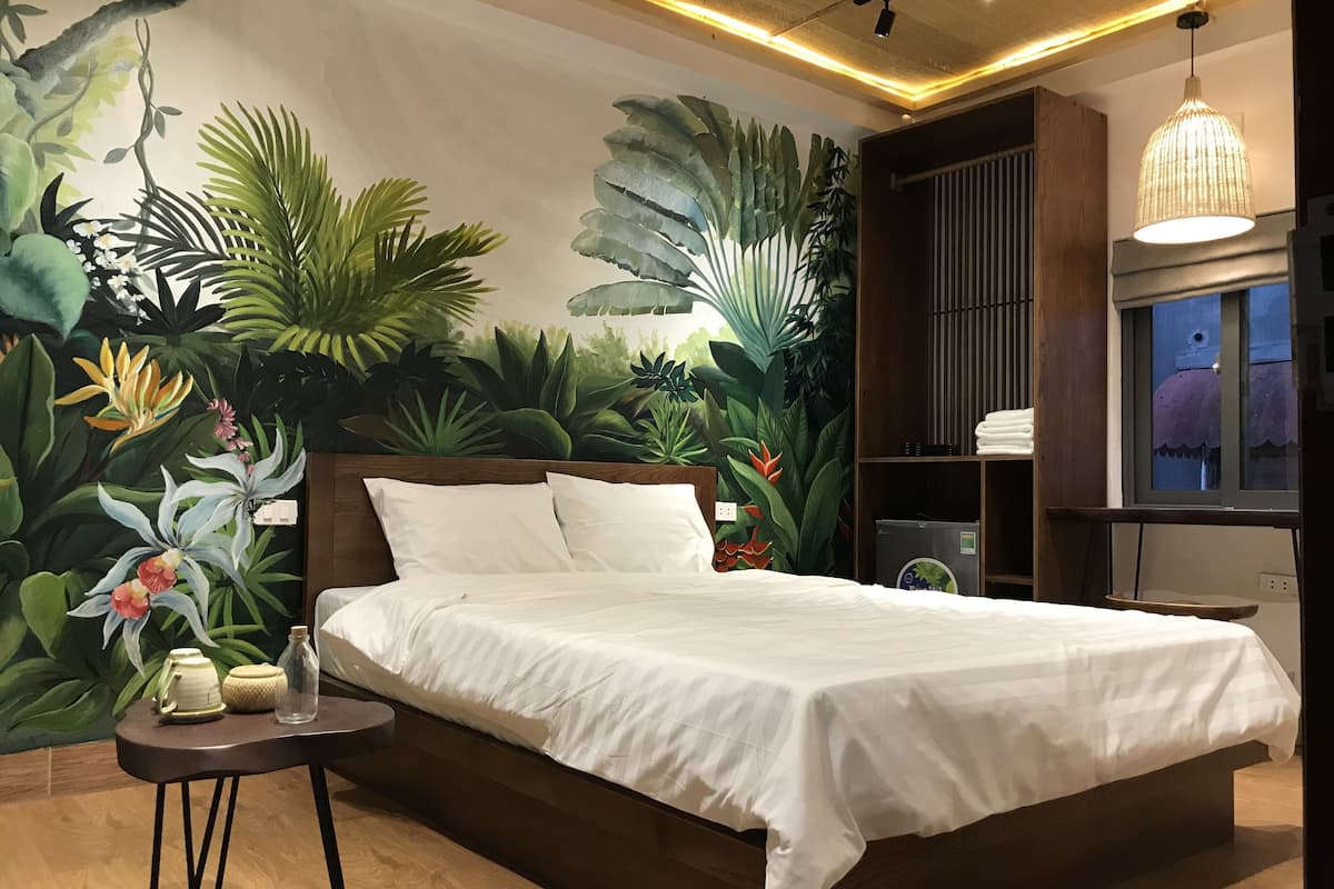 Un dormitorio con una cama grande, una mesita de noche, un mural de plantas tropicales, una lámpara colgante y una ventana con vistas.