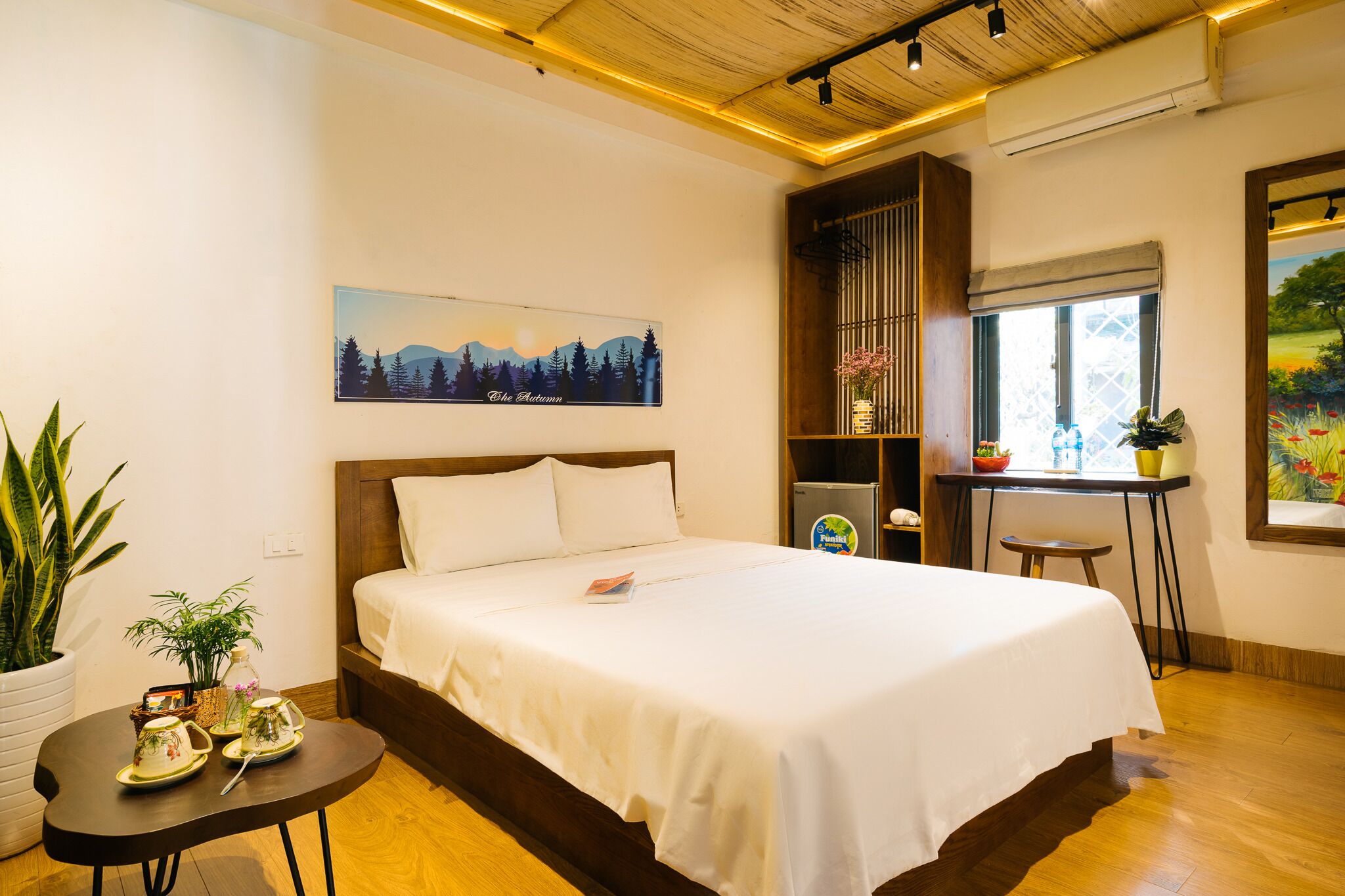 Habitación de hotel ordenada con cama, una mesita con objetos decorativos, una ventana con vistas y un cuadro en la pared.