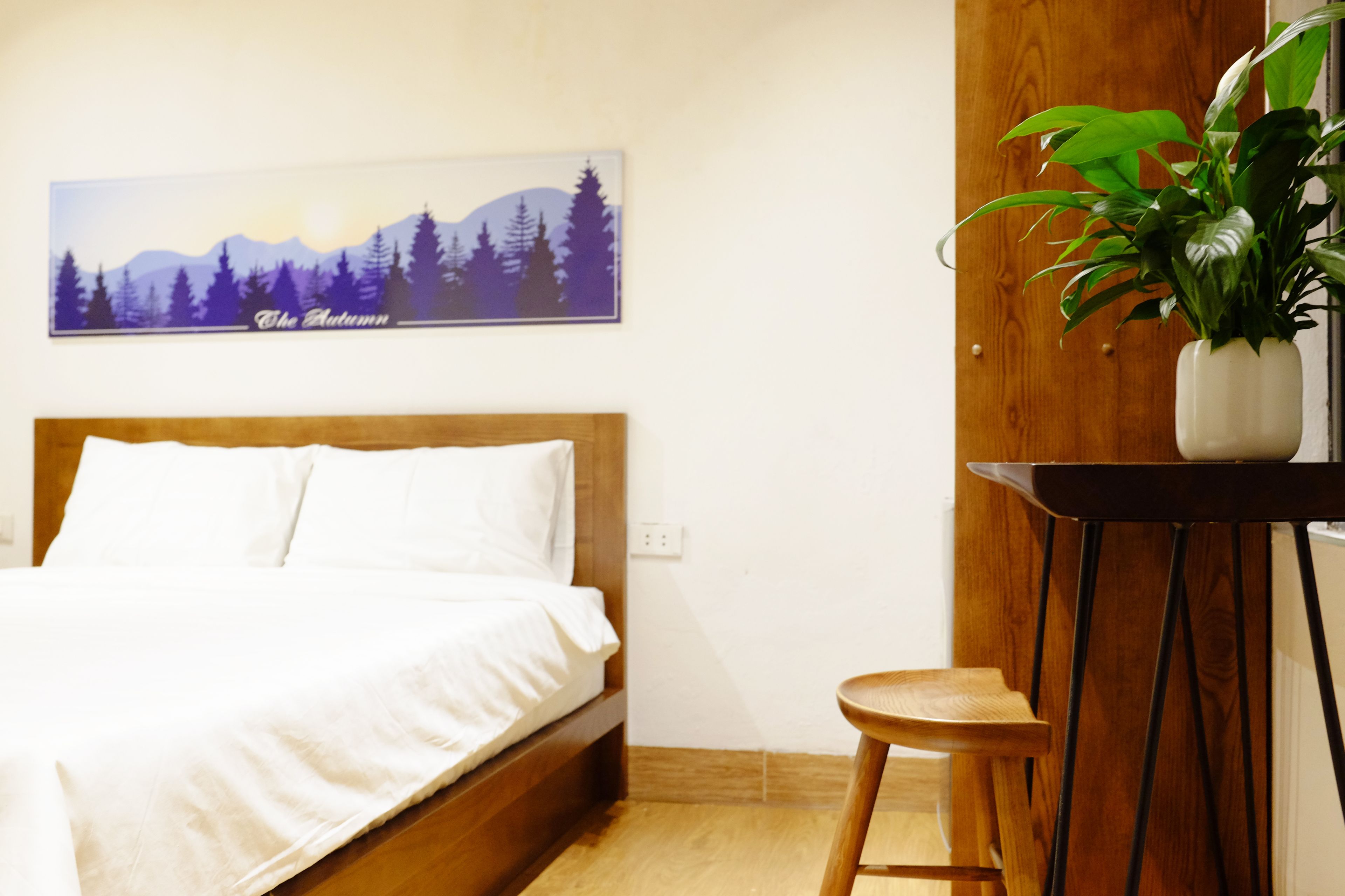 Une chambre à coucher avec un lit, une tête de lit en bois, un tableau accroché au mur, une plante en pot et un petit tabouret en bois.