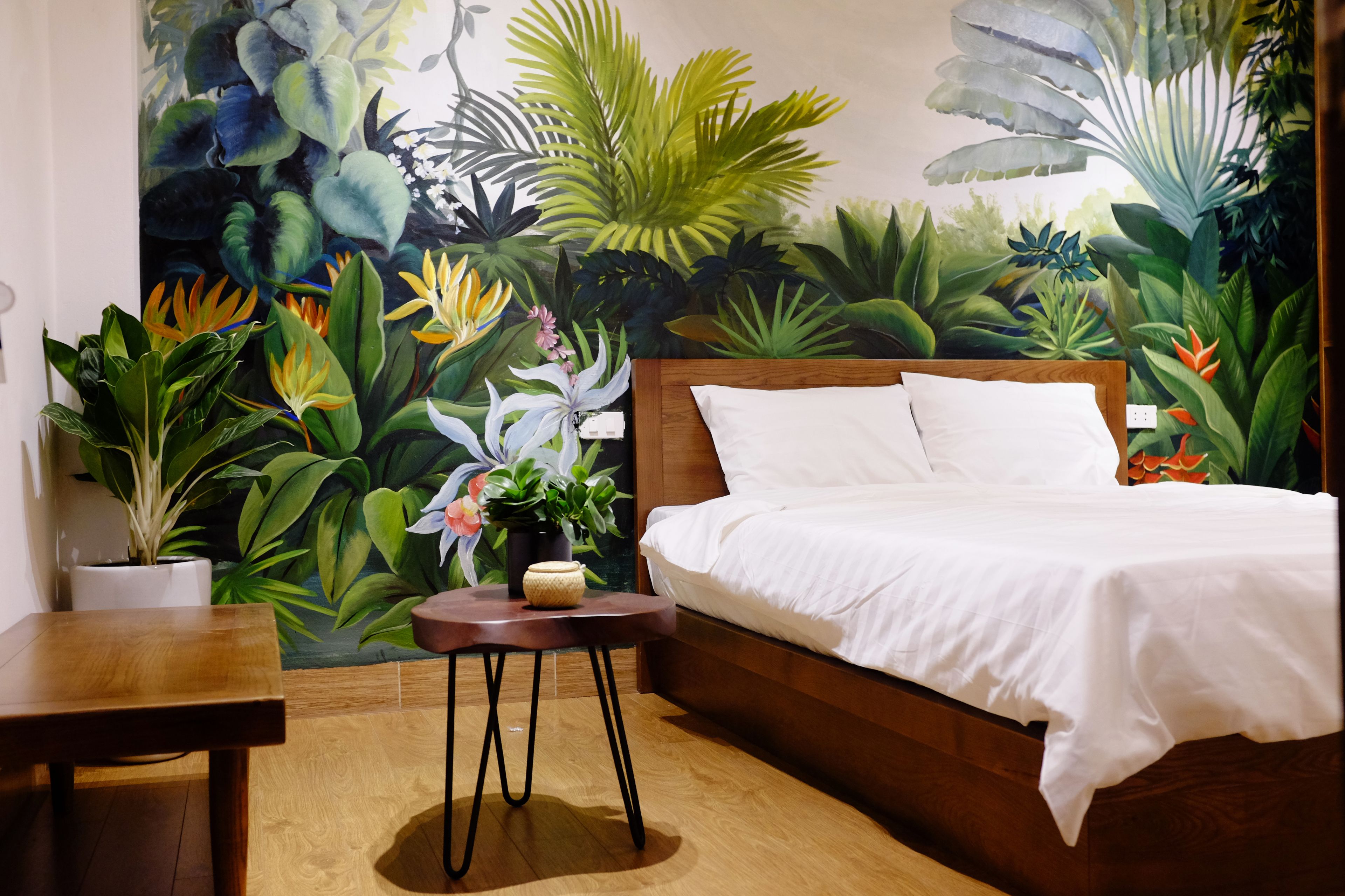 A bedroom with a large mural of tropical plants on the wall, a wooden bed, and a small round table with a plant.