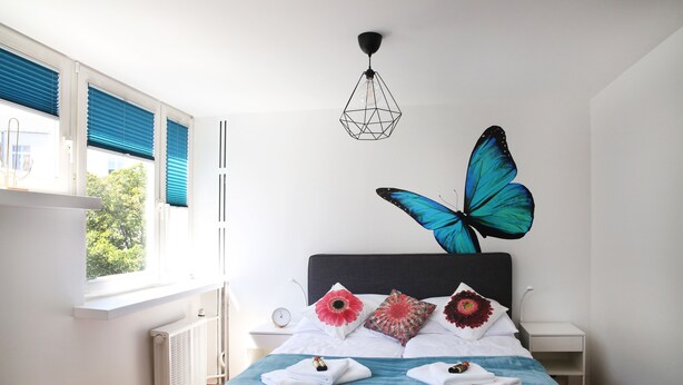 A bedroom with a bed, a large butterfly wall art, a window with blinds, a bedside table with a clock, and a radiator. - Plac Teatralny - Imaginea City Apartments (Warsaw)