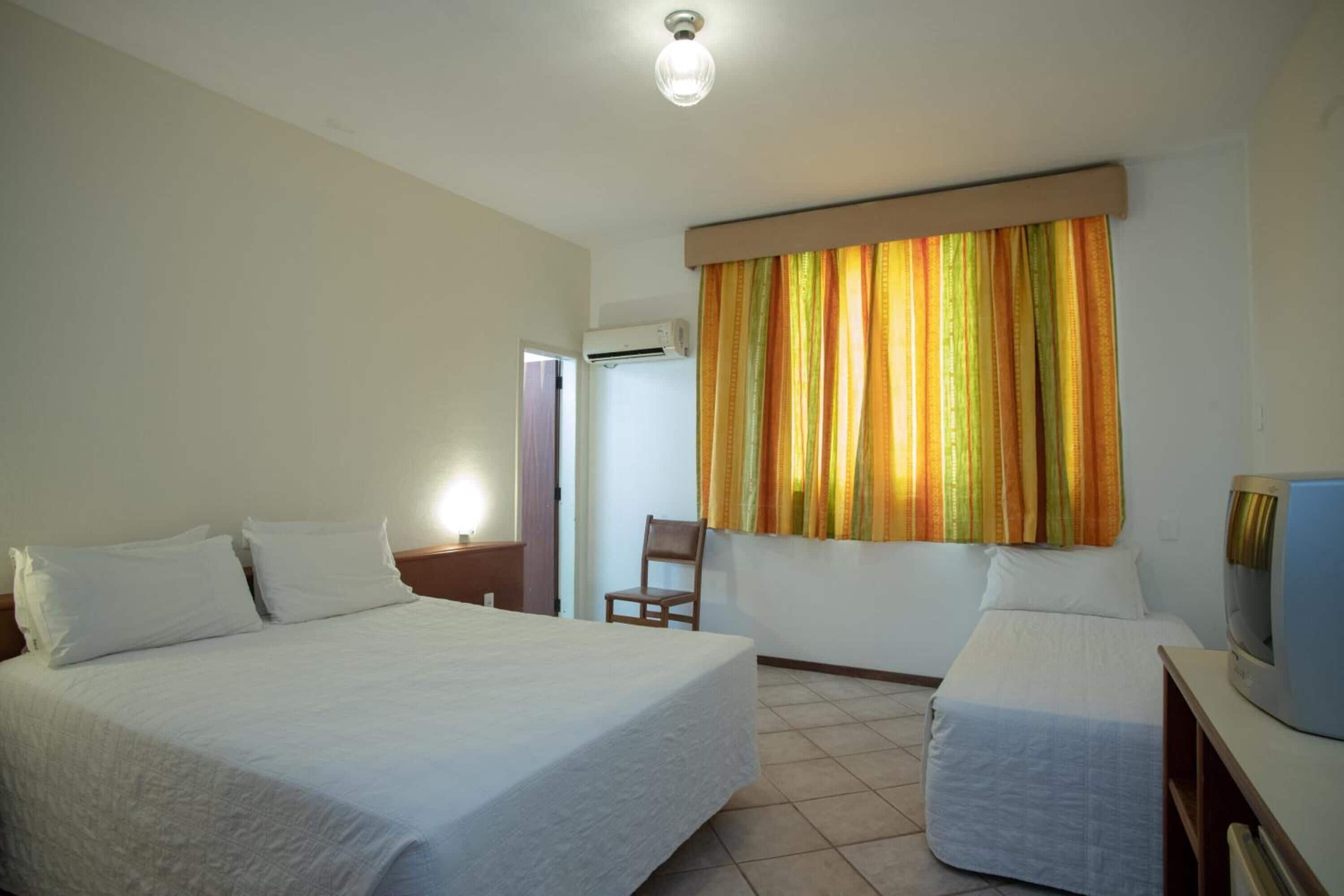 A hotel room with a large bed, a smaller bed, a TV, and an air conditioning unit.