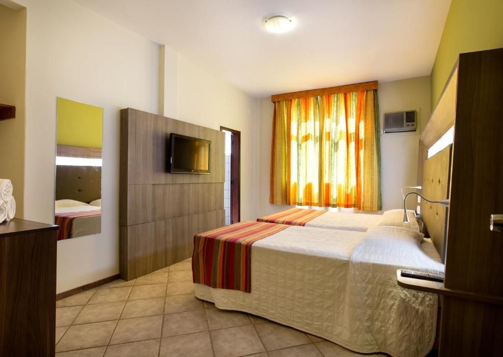 A hotel room with a bed, a television, a mirror, and a window with curtains.
