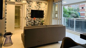 Una sala de estar moderna con un sofá, un televisor de pantalla plana montado en una pared con diseño geométrico, una puerta de cristal que da a un balcón y un comedor con mesa y sillas.