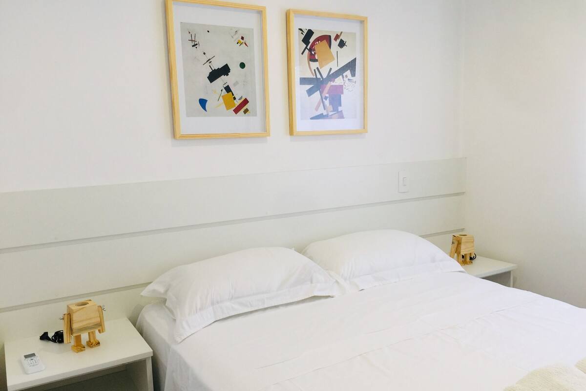 A hotel room with a bed, white bedding, two framed abstract artworks on the wall, a bedside table with a wooden figurine, and a small shelf with a white object.