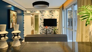 Un salón moderno con una lámpara de techo negra, un televisor de pantalla plana montado en una pared con un diseño geométrico, y una planta grande a la derecha.