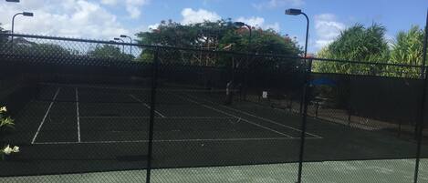 Una cancha de tenis rodeada por una cerca de malla metálica, con un cielo despejado y árboles al fondo.
